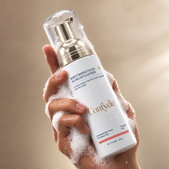 A hand wrapped in white cleansing foam holds a bottle of Confyde Moisturizing Foam To-Beam Cleanser. The bottle is white with a metallic gold pump and clear cap, set against a soft beige background.